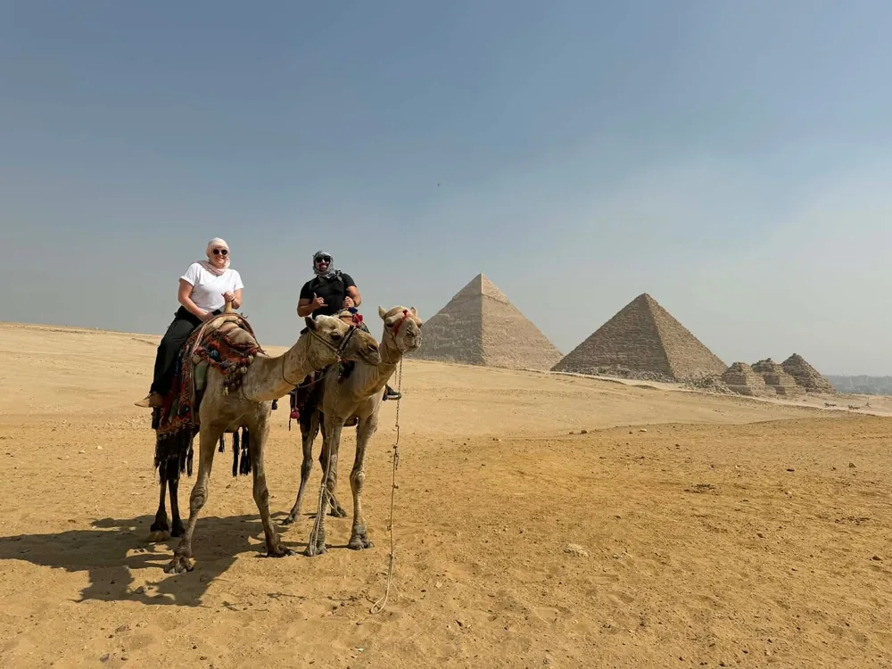 Two riders sitting on camels on the sand, with the pyramids of Khafre and Menkaure and the small queens' pyramids behind them