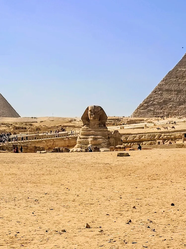 The Great Sphinx seen from the sand in front, between two pyramids, with visitors on the terrace to the left